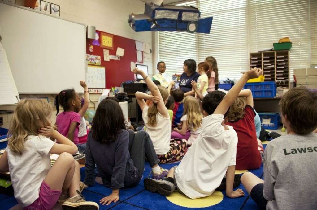 Image shows a classroom with a class of children with a teaching assistant